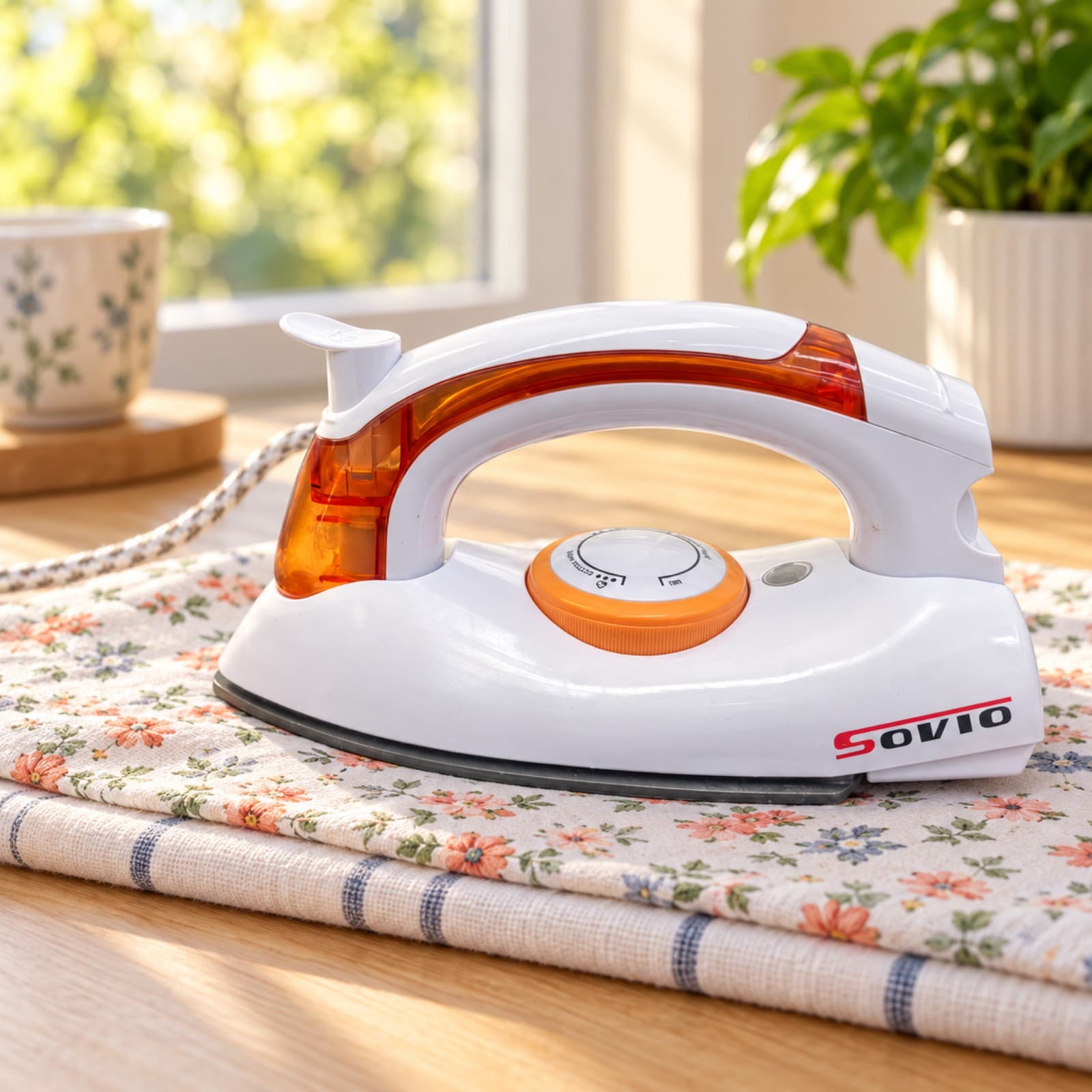 Plancha de viaje Sovio S-258B naranja sobre un paño floral en una mesa de madera junto a una ventana (imagen ilustrativa) — miniatura 7